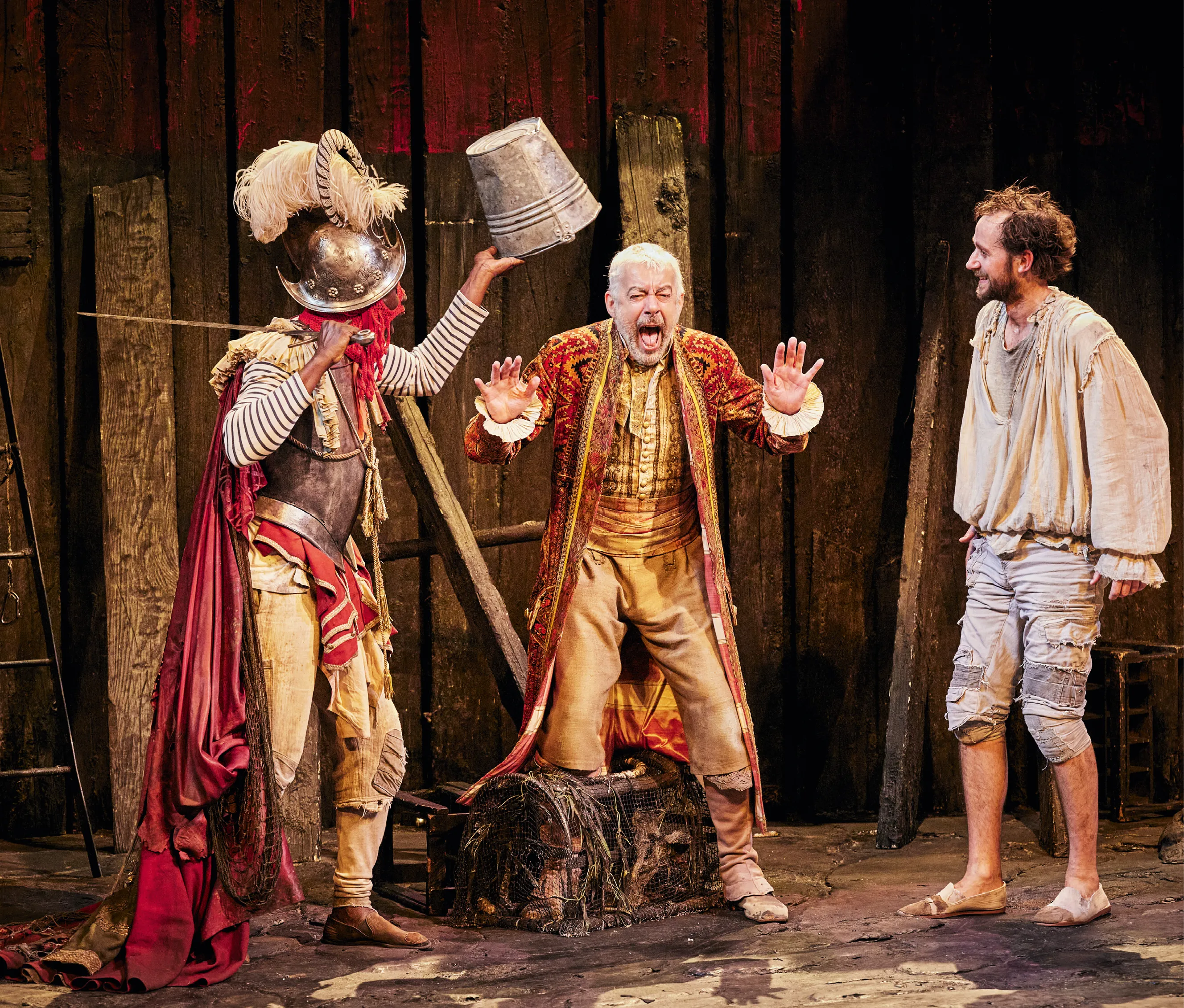 Photographie d'une scène de théâtre issue des Fourberies de Scapin, mise en scène par Denis Podalydès à la Comédie-Française en 2017. On y voit trois personnages : Silvestre (en costume de soldat avec un seau), Géronte (en costume noble, terrifié) et Scapin (en vêtements simples, souriant).