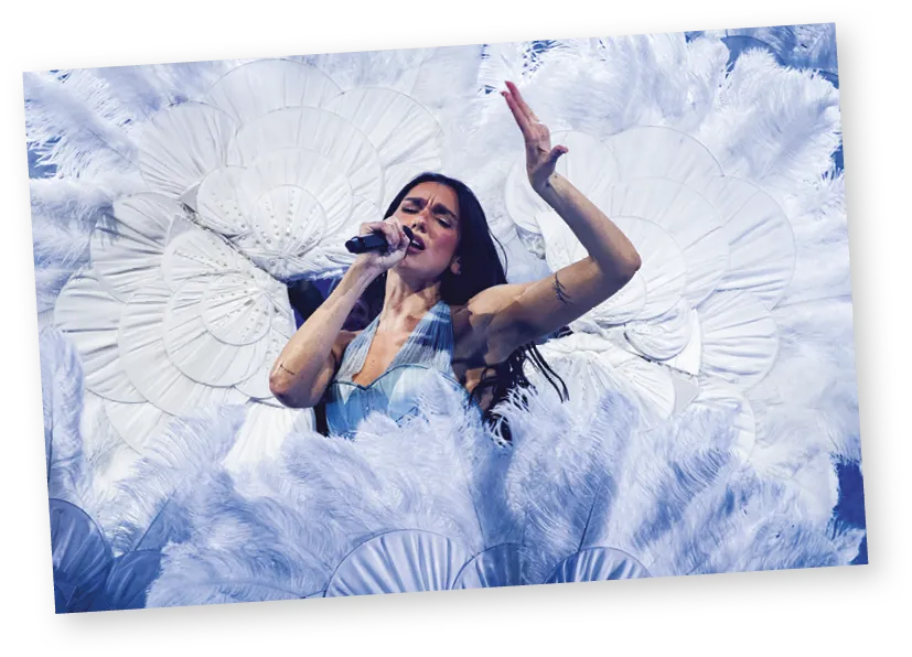 Dua Lipa performing on stage surrounded by large white feathers.