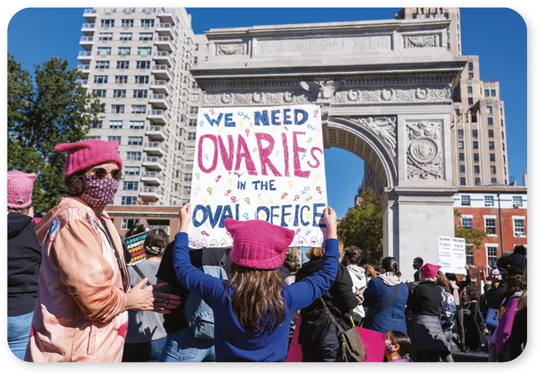 Protesters holding a sign reading 'We need ovaries in the Oval Office' in front of a large arch monument.