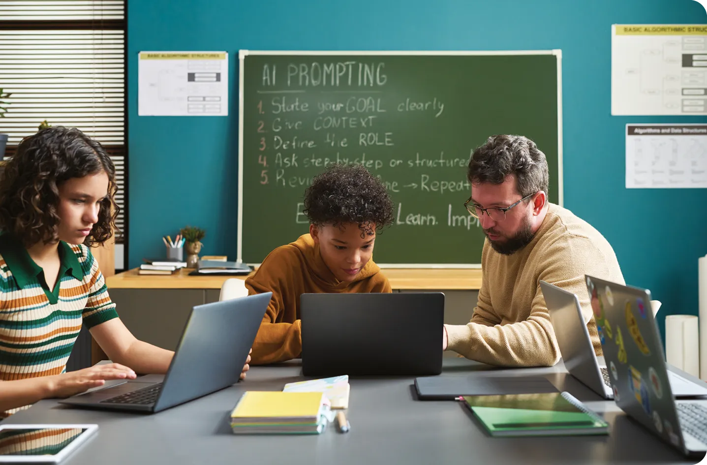 Homme aidant un adolescent et une adolescente travaillant sur des ordinateurs portables dans une salle de classe, tableau noir avec IA demandant des instructions visibles en arrière-plan.