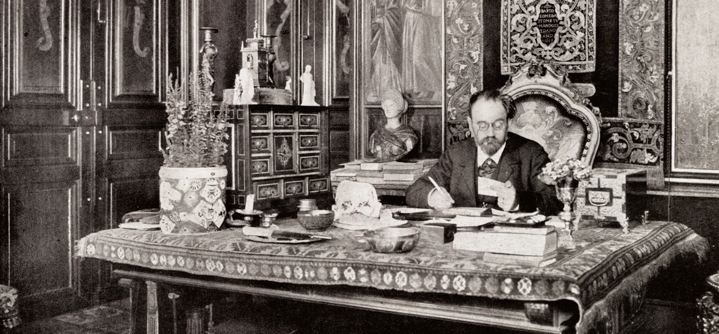 Photographie : Émile Zola, écrivain, assis à son grand bureau décoré, écrit. Bureau richement orné, ambiance feutrée.