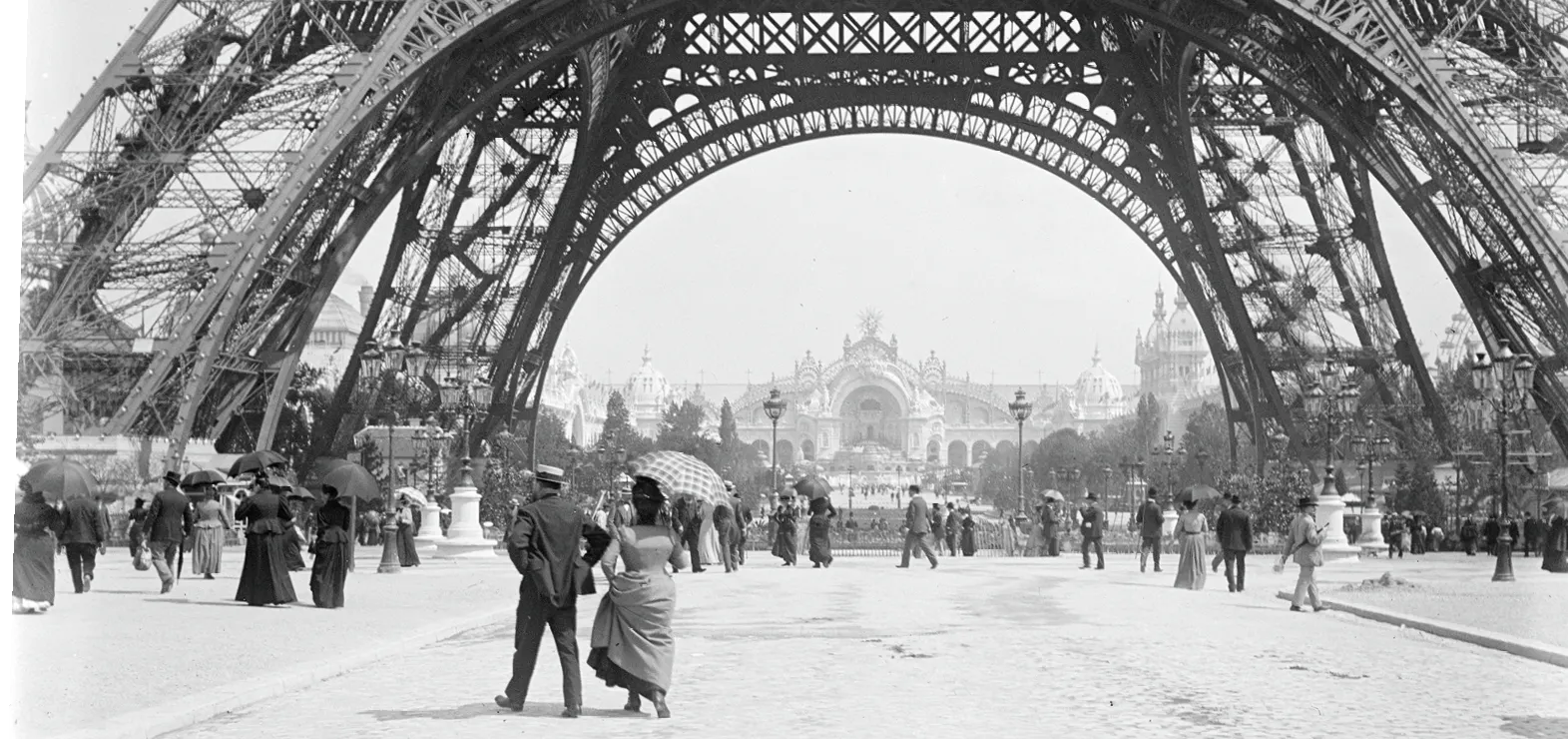Photographie ancienne : couple se promenant sous la Tour Eiffel, visiteurs avec parapluies, Exposition universelle de Paris en arrière-plan.