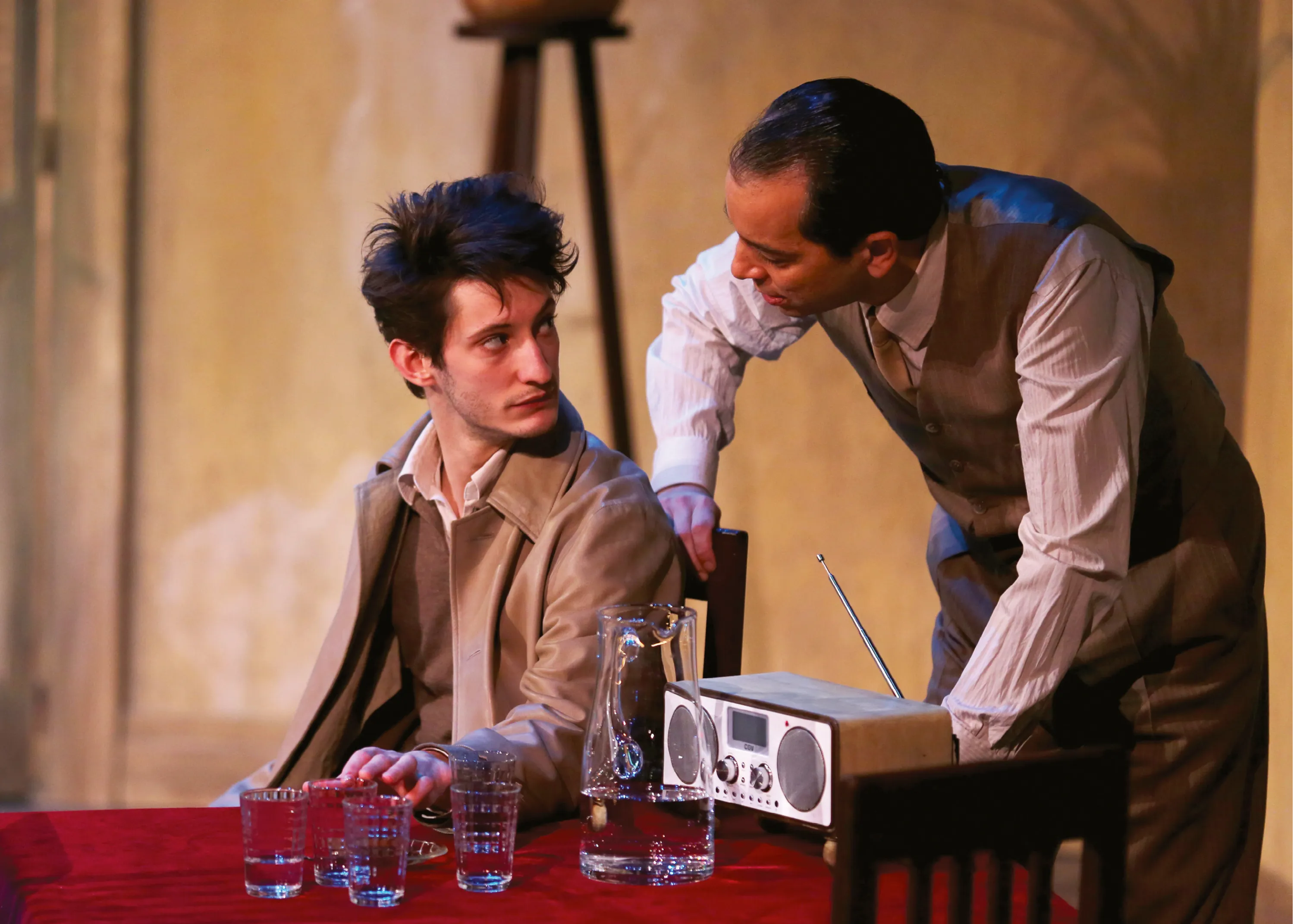Photographie de scène de théâtre : Pierre Niney et Éric Génovèse dialoguent autour d'une table. Ambiance feutrée.