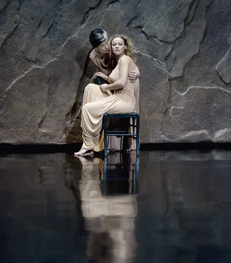 Photographie de scène théâtrale : Phèdre et OEnone, en robes sable, interagissent sur scène sombre. OEnone est assise.
