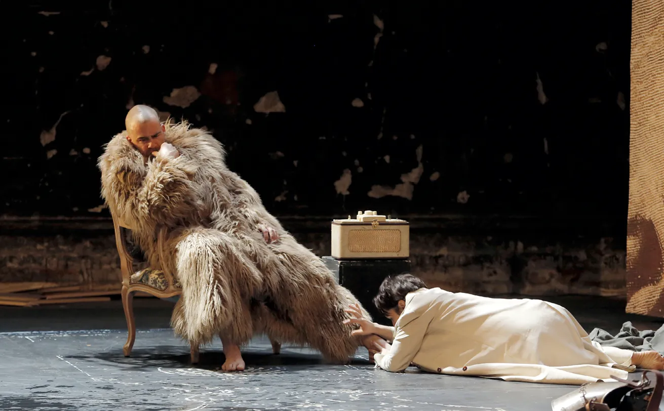 Photographie de scène théâtrale : Phèdre (Cécile Garcia Fogel), longue fourrure beige, assise, Thésée (Olivier Werner) à ses pieds.