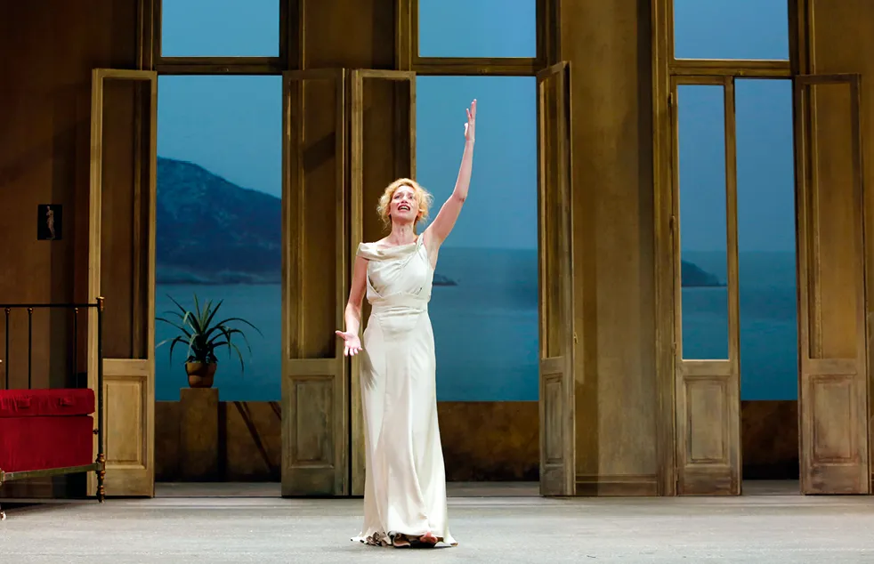 Photographie de scène de théâtre : Elsa Lepoivre, actrice en robe blanche, bras levé, sur fond marin et montagneux. Comédie-Française (2013).
