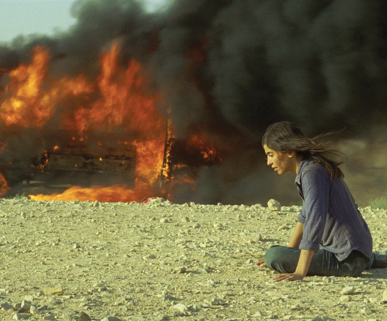 Photogramme : femme assise, véhicule brûlant en arrière-plan. Incendies.