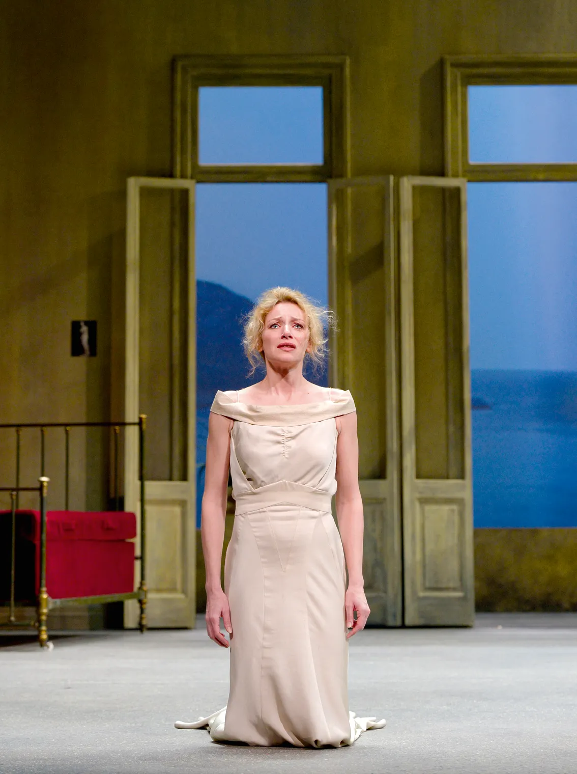 Photographie : Elsa Lepoivre, dans le rôle de Phèdre, agenouillée, robe beige, regarde par une fenêtre, vue mer.