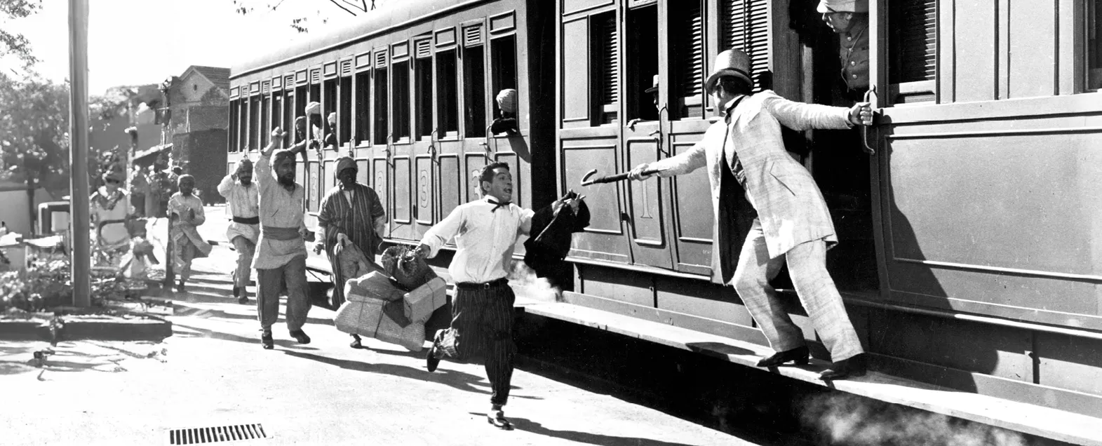 Photographie noir et blanc du film &quotLe tour du monde en 80 jours" (1956). Cantinflas et David Niven, poursuivis par une foule, embarquent dans un train en marche.