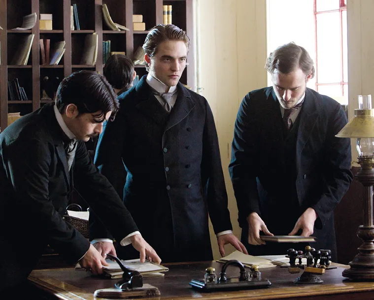 Capture du film Bel Ami (2012) : Robert Pattinson et deux autres hommes en costume d'époque examinent des documents sur un bureau.
