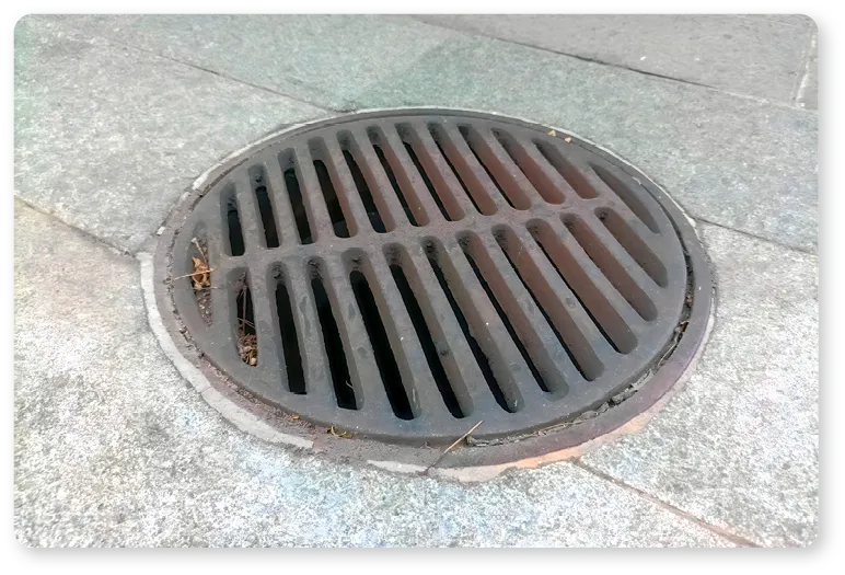 Plaque d'égout circulaire avec grille métallique composée de barres parallèles, posée sur pavés en pierre. Exemple d'élément géométrique dans l'environnement urbain.