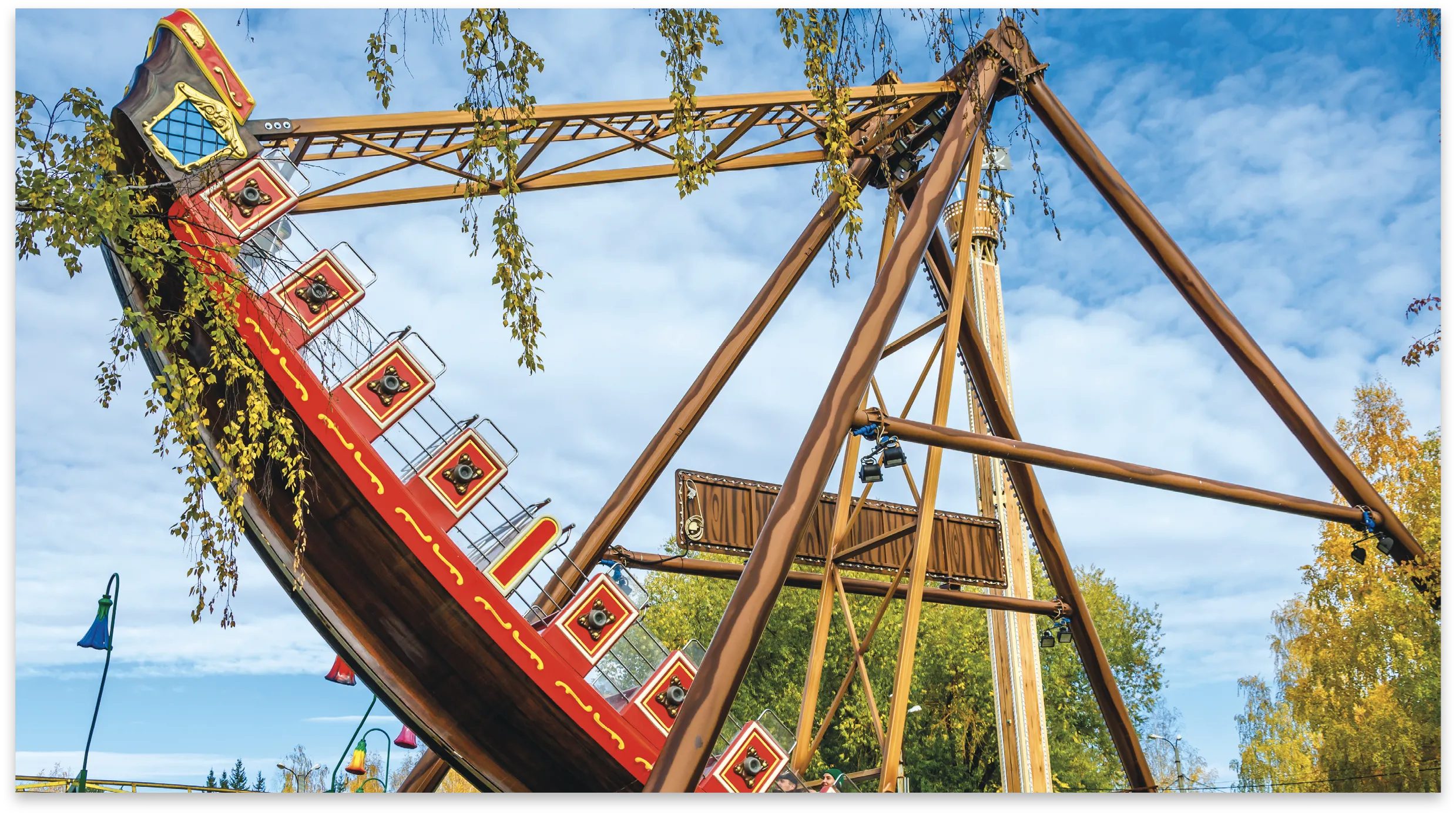 Photographie d'une attraction de parc d'attractions, une nacelle en forme de bateau pirate, inclinée, vue de dessous. Structure en bois, ciel bleu et arbres automnaux.