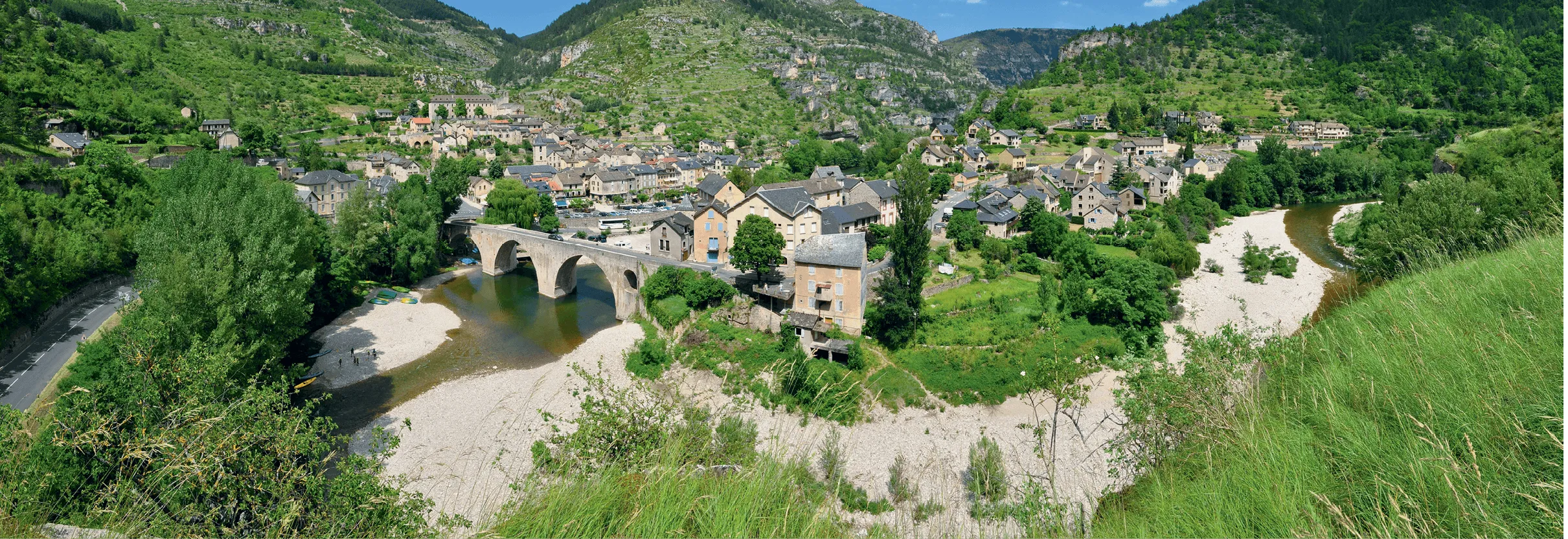 Photo aérienne d'un village perché traversé par une rivière et un pont de pierre. Architecture en pierre, végétation luxuriante.