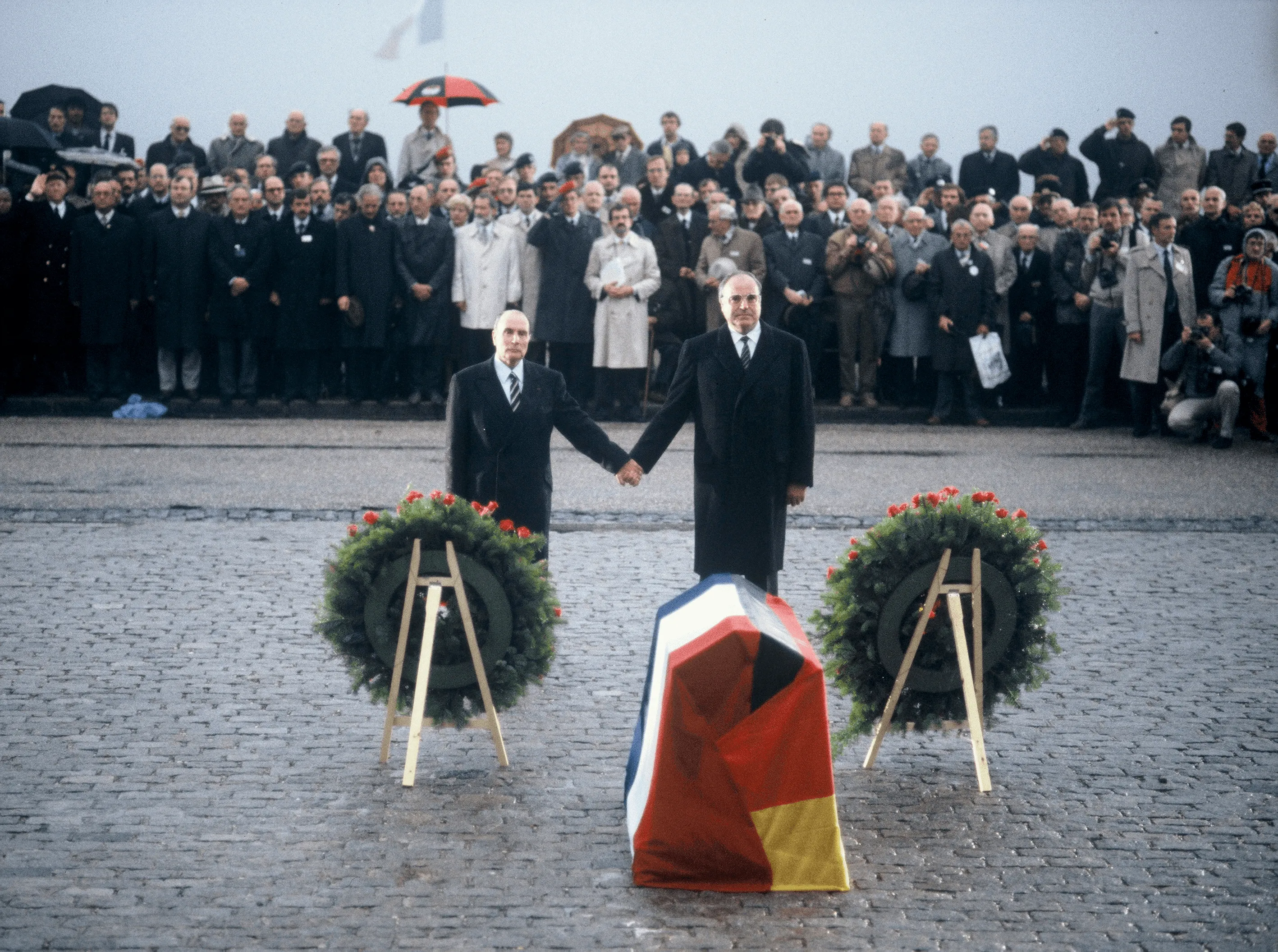 Photographie de François Mitterrand et Helmut Kohl se tenant la main devant un cercueil drapé des drapeaux français et allemand.