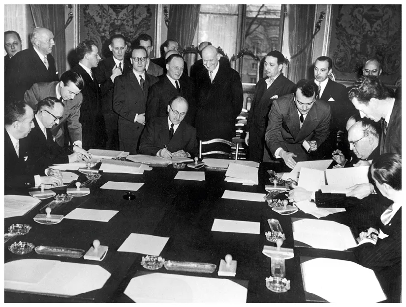 Photographie historique en noir et blanc du traité de Paris (1951), montrant de nombreux hommes en costume signant des documents importants autour d'une grande table.