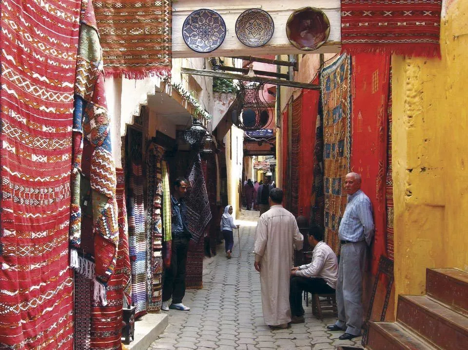 Photographie d'une ruelle animée de la médina, bordée de tapisseries colorées et d'artisans. Ambiance commerçante et chaleureuse.