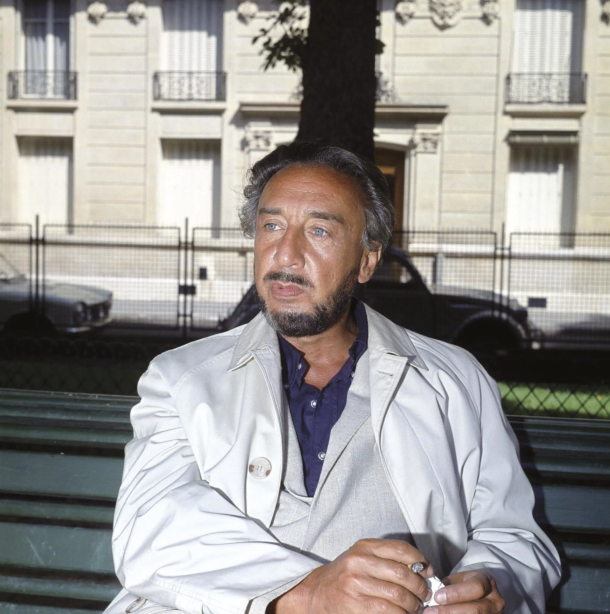 Photographie de Romain Gary assis sur un banc à Paris, vêtu d'un imperméable beige. Il tient une cigarette.