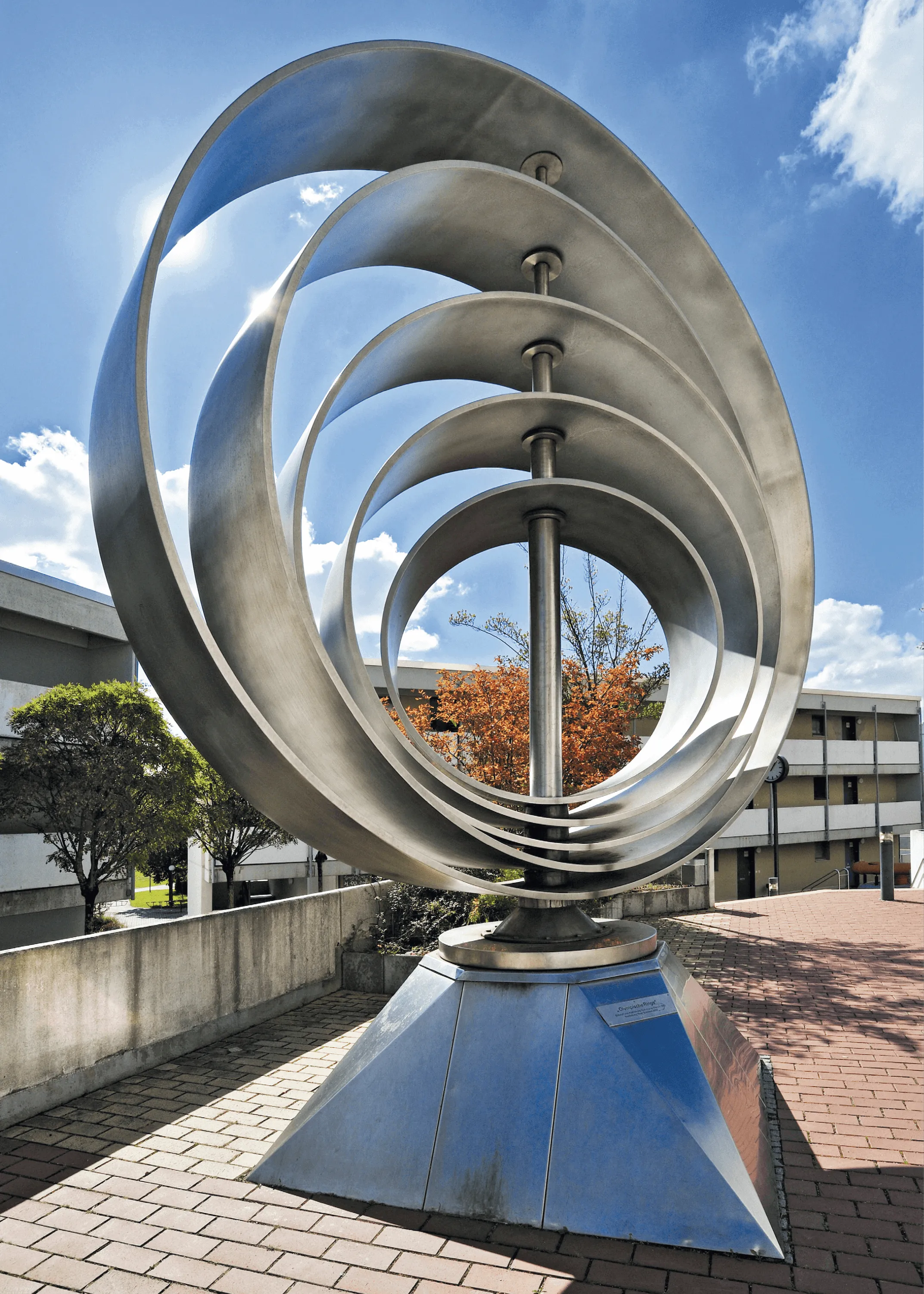 Sculpture métallique : anneaux entrelacés, formant un cercle, sur un piédestal. Extérieur, ciel bleu.