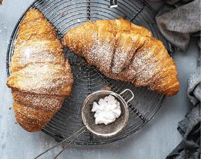 Photographie aérienne de deux croissants dorés, saupoudrés de sucre glace, reposant sur une grille métallique. Un tamis contenant du sucre glace est visible à côté.