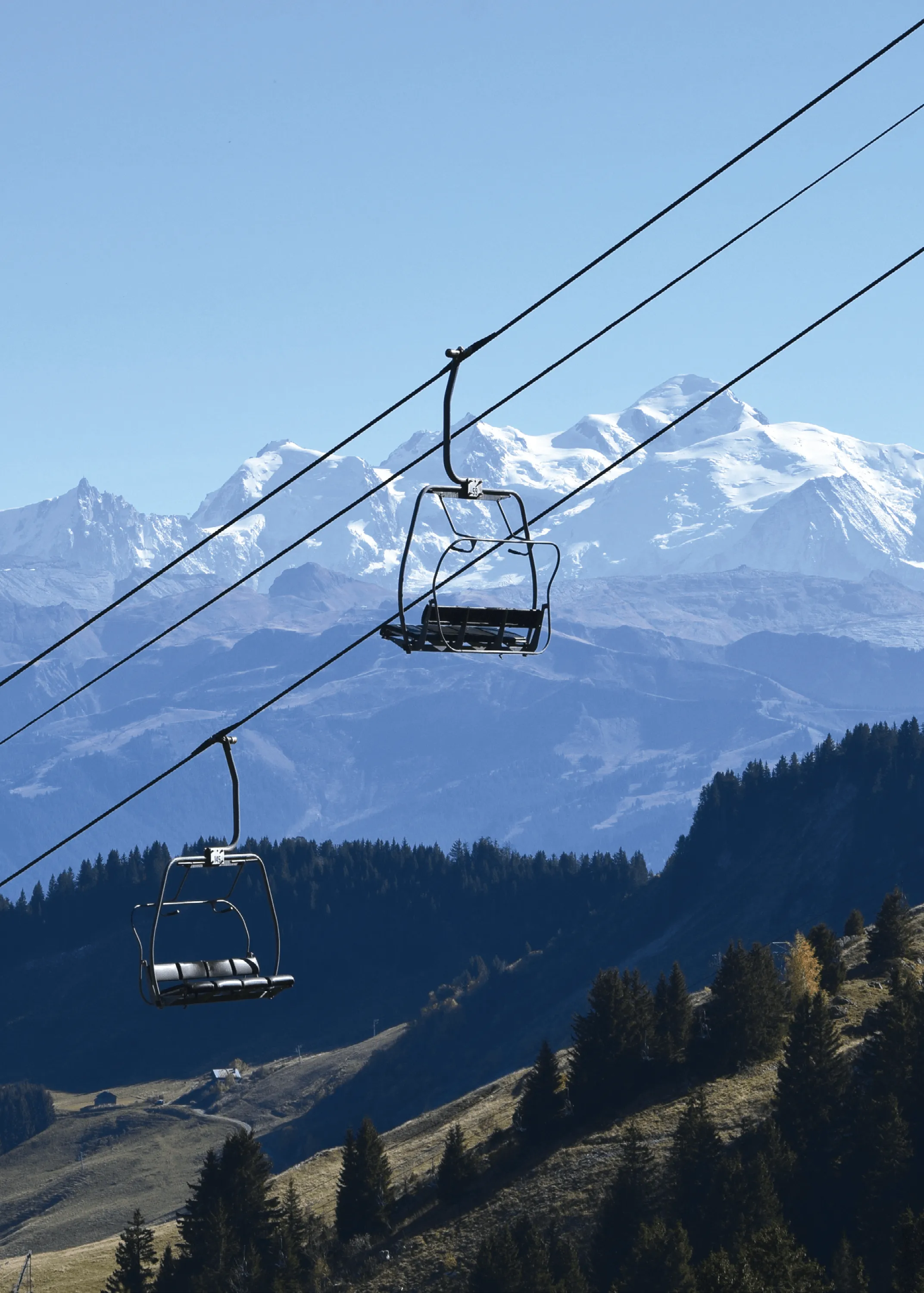 Photographie de deux télésièges vides au-dessus d'une montagne enneigée.  Les montagnes et forêts environnantes sont visibles par temps clair.