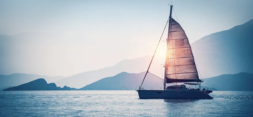 Photographie d'un catamaran naviguant au coucher du soleil, au large de montagnes bleutées.