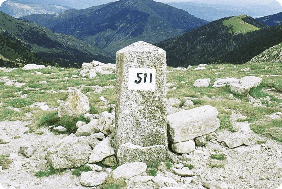 Photographie d'une borne frontière numéro 511 en montagne. La borne en pierre est entourée de rochers et d'herbe. En arrière-plan, des montagnes verdoyantes.