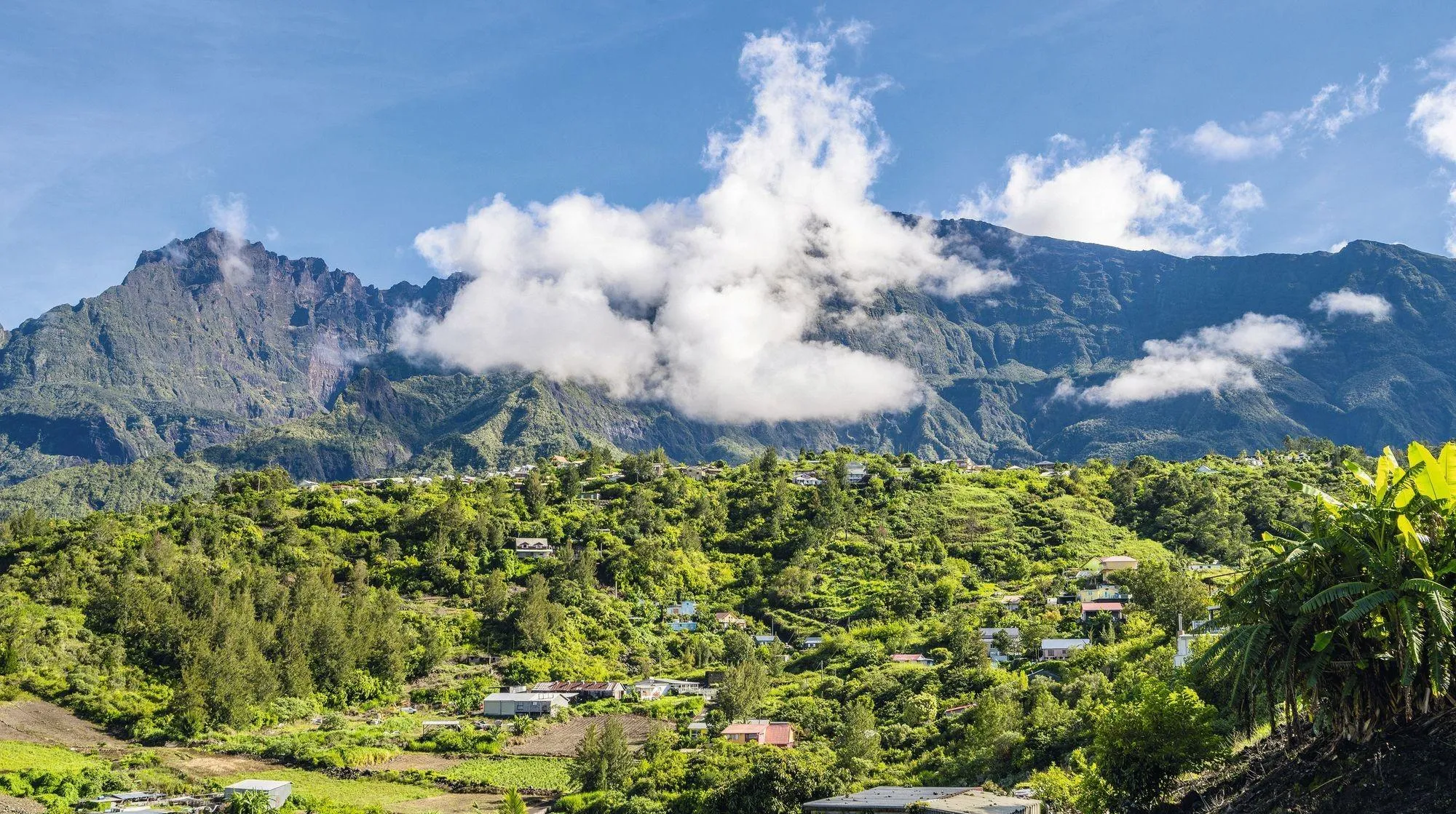 Photographie du cirque de Cilaos à La Réunion. Des maisons sont nichées dans la végétation luxuriante au pied de montagnes imposantes partiellement voilées par les nuages.