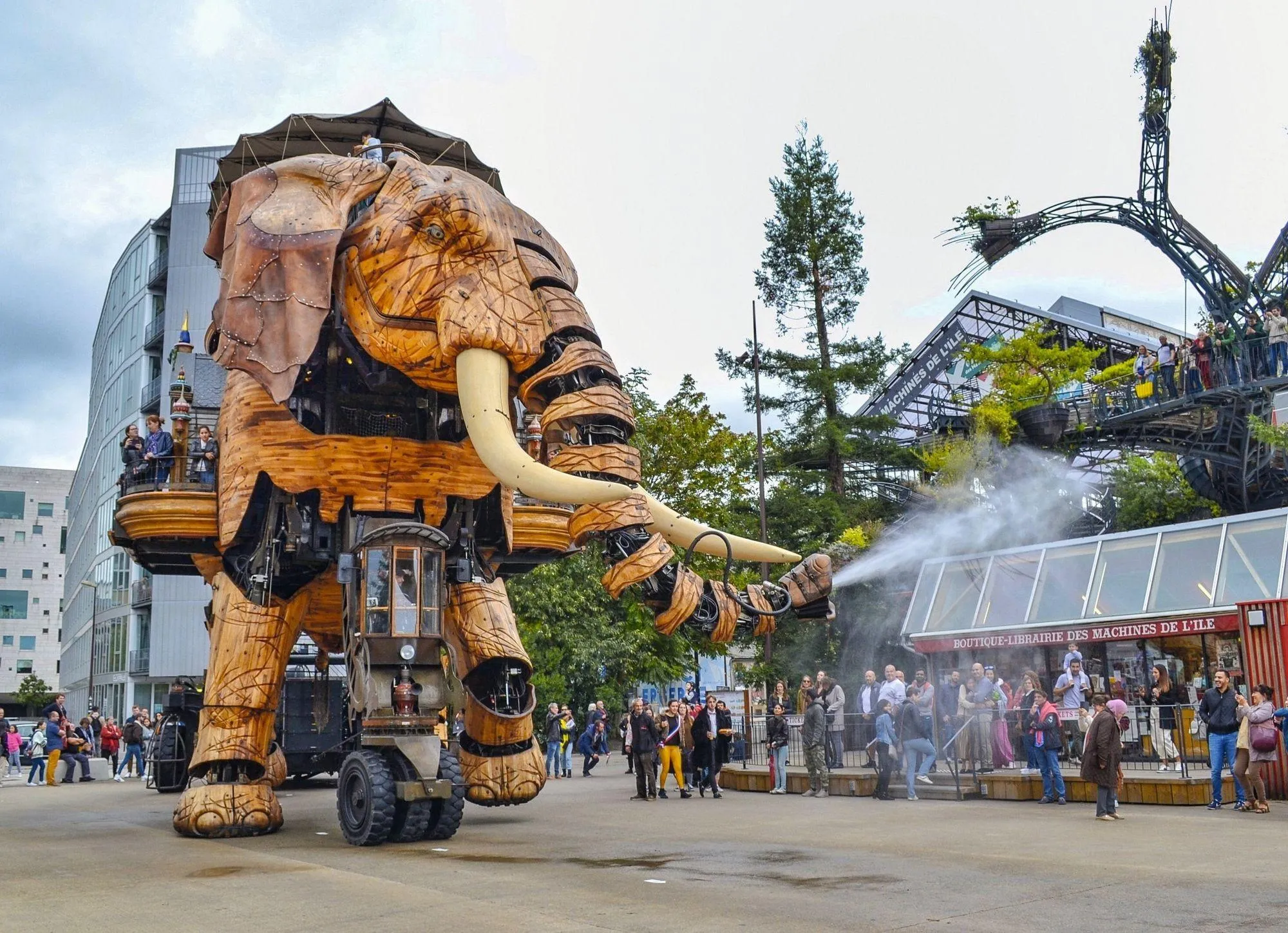 Photographie d'un éléphant mécanique géant à Nantes, projet des Machines de l'Île, arrosant les visiteurs.  Structure en bois, détails mécaniques visibles.