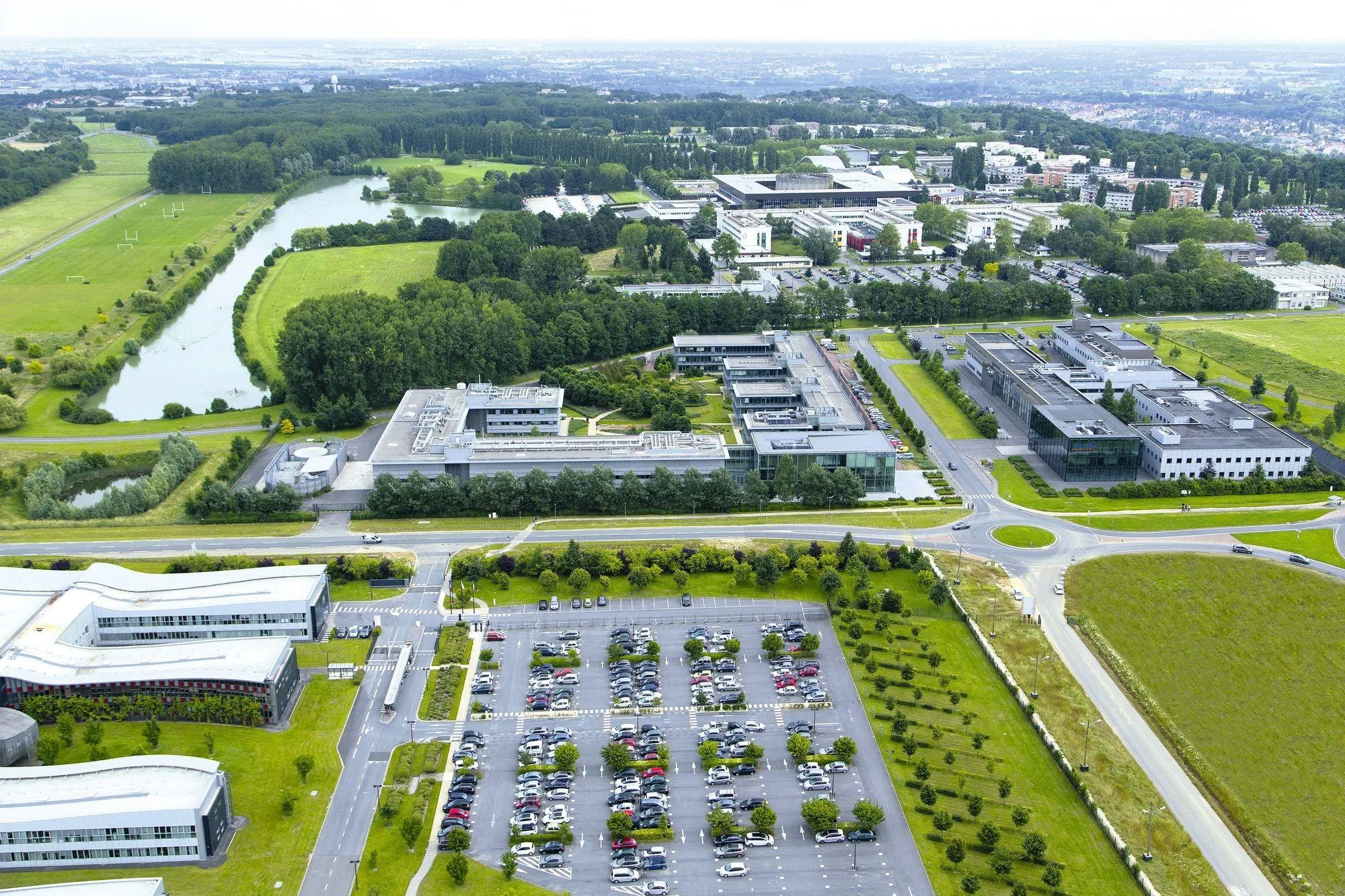 Photo aérienne du campus de Paris-Saclay, montrant de nombreux bâtiments modernes, un grand parking et un environnement verdoyant.