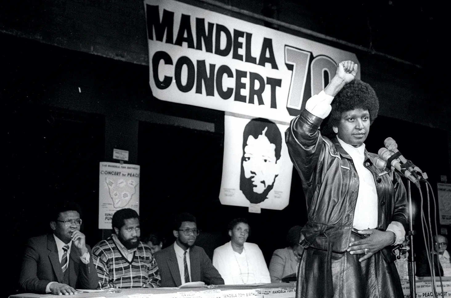 Photographie : Winnie Mandela prend la parole lors d'un concert pour le 70e anniversaire de Nelson Mandela. Elle porte un blouson en cuir et lève le poing.