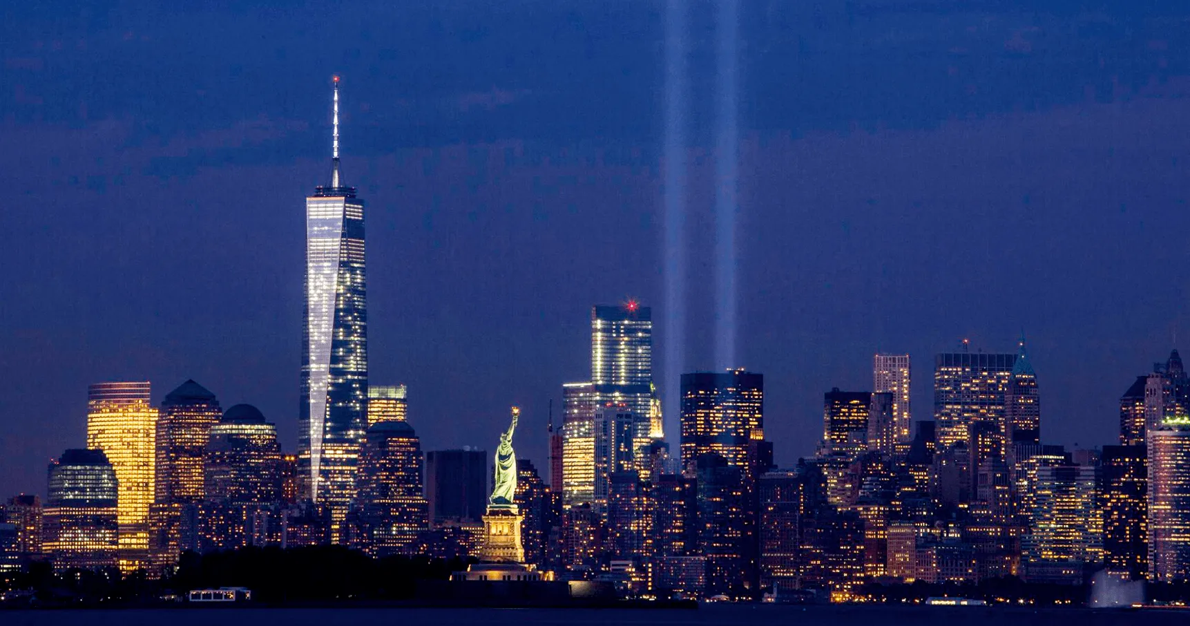 Photographie nocturne de la skyline de New York illuminée, incluant la Statue de la Liberté et le One World Trade Center, avec des faisceaux lumineux commémoratifs.