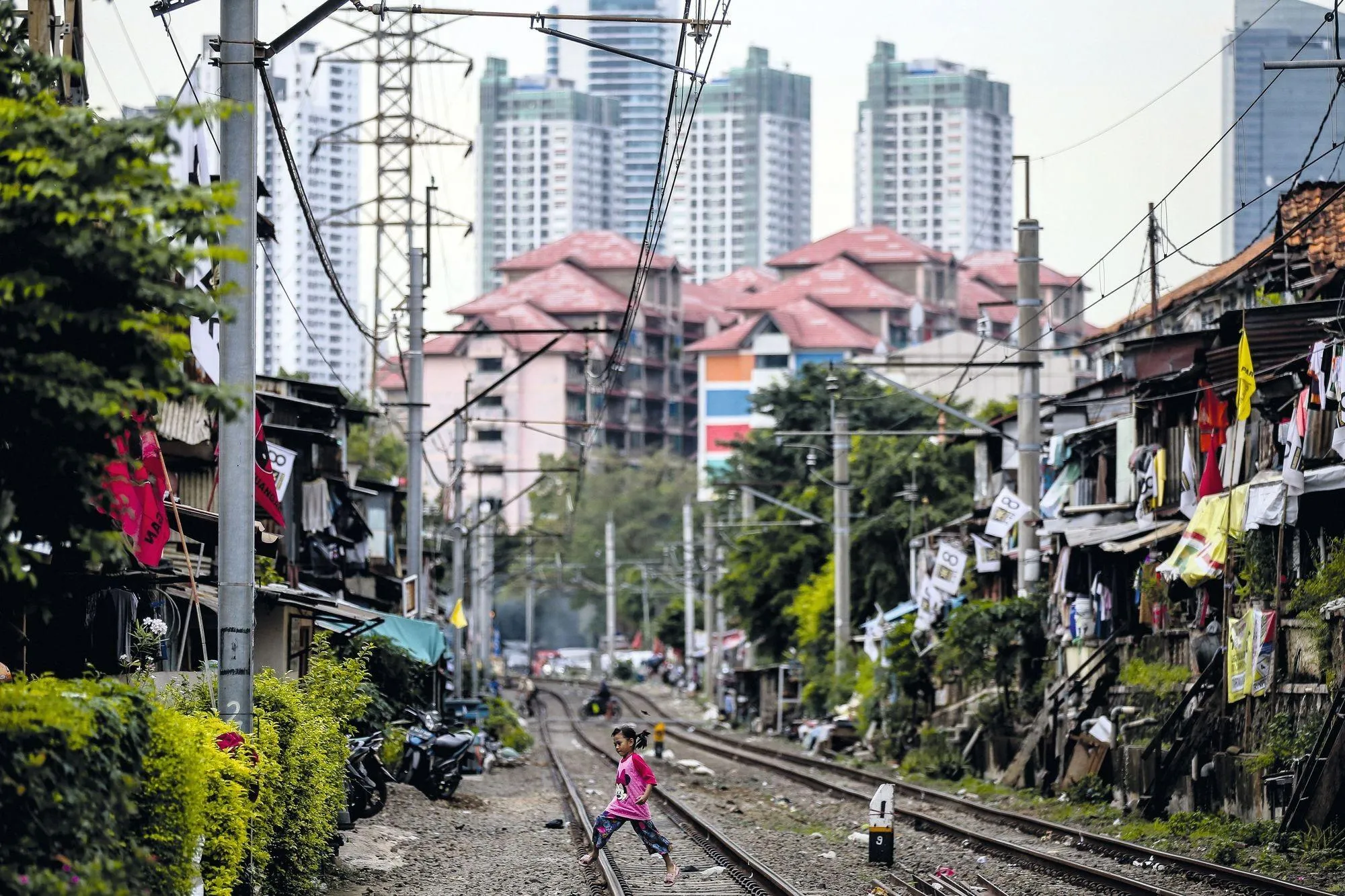 Photographie de Jakarta montrant une fillette marchant sur des voies ferrées, entre des habitations précaires et des immeubles modernes.  Inégalités sociales.