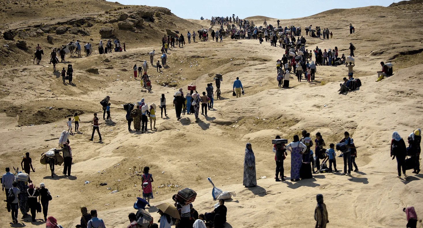 Photographie d'un grand groupe de réfugiés syriens marchant dans un paysage désertique aride. Ils portent des bagages et certains animaux les accompagnent.