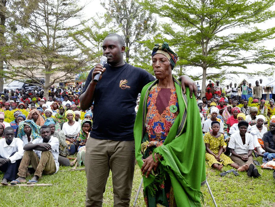 Photographie : Commémoration du génocide rwandais. Un homme prend la parole au micro, une femme âgée l'écoute, entourées de nombreuses personnes.