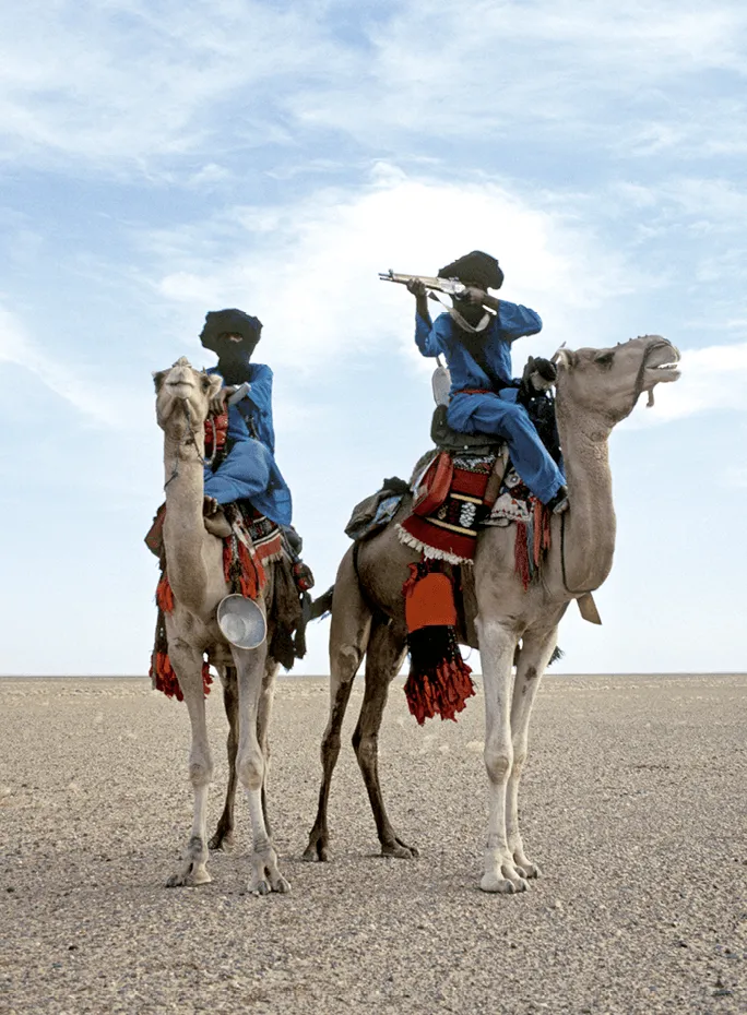 Photographie de Frans Lemmens (2000) : deux Touaregs en bleu, armés, à dos de chameau dans le désert.