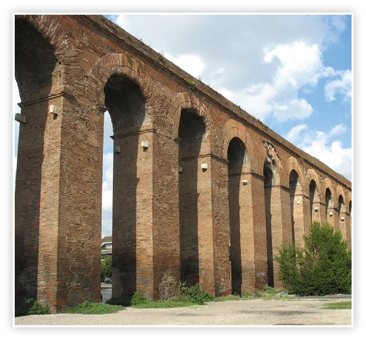 Photographie d'un aqueduc romain antique en briques, montrant ses arches et ses piliers sous un ciel bleu.