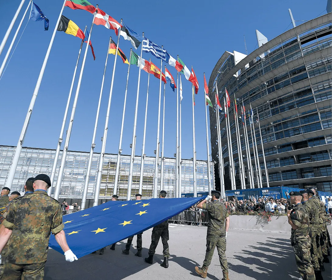 Photographie de soldats déployant le drapeau européen devant le Parlement européen.  Plusieurs drapeaux nationaux européens sont visibles en arrière-plan.