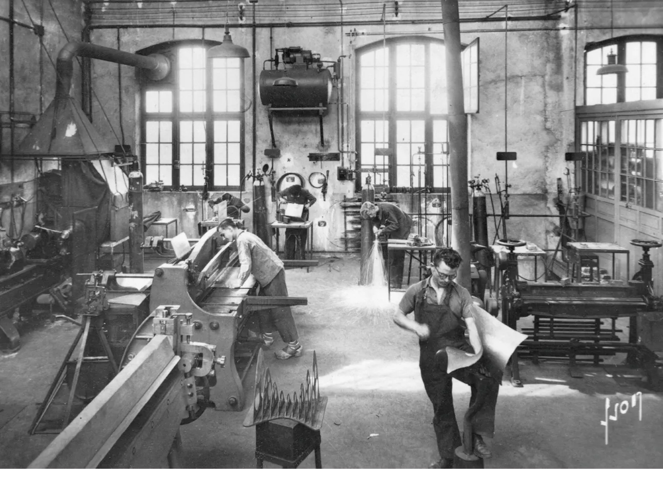 Photographie en noir et blanc d'un atelier d'étudiants en école professionnelle, travaillant sur des machines industrielles.