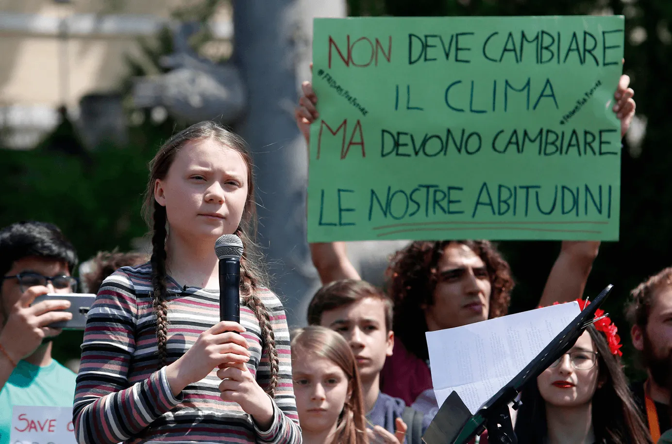 Photographie de Greta Thunberg, jeune activiste climat, s'exprimant au micro lors d'une manifestation.  Elle tient un micro et derrière elle, des jeunes manifestants brandissent des pancartes.