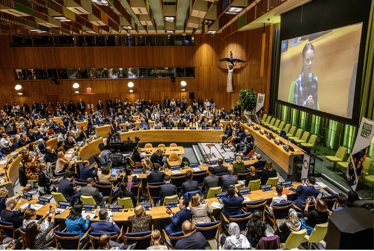 Photographie d'une assemblée plénière à l'ONU où une jeune femme prend la parole sur écran, suivie par une assemblée attentive.