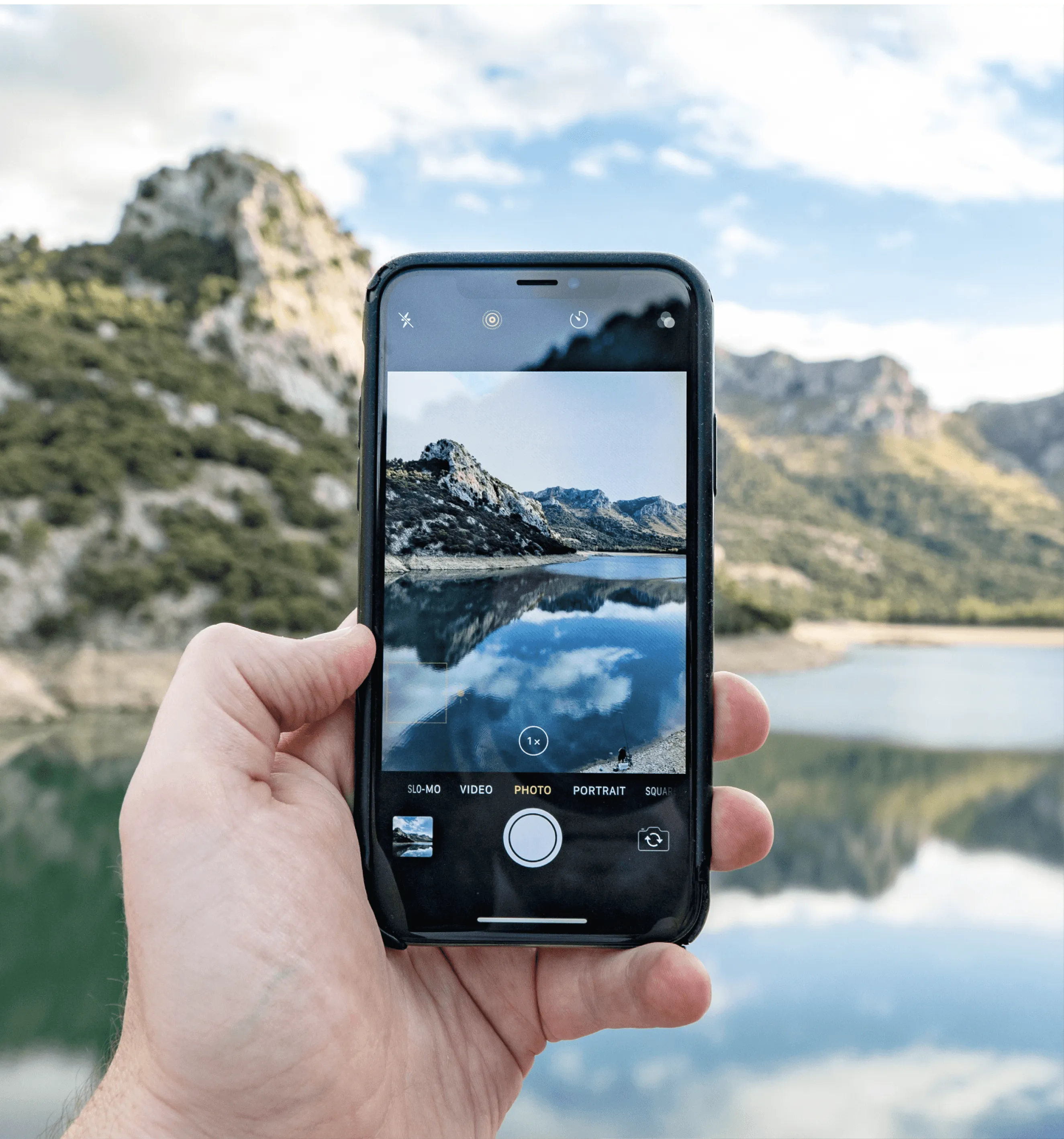 Photographie d'un smartphone affichant une photo de paysage montagneux et lac. Main tenant le téléphone, reflet du ciel dans l'eau.