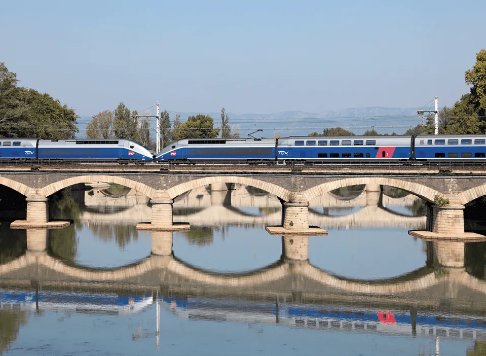 Photographie de deux TGV traversant un pont de pierre au-dessus d'une rivière. Reflet du train et du pont dans l'eau.