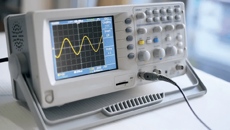 Photographie d'un oscilloscope affichant une onde sinusoïdale. L'appareil est blanc et gris, avec de nombreux boutons et un écran LCD. Un câble est branché.