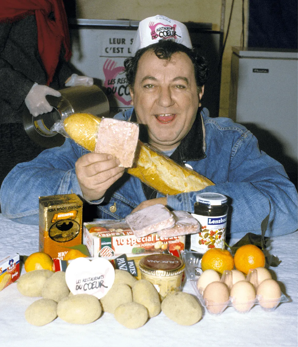 L'humoriste Coluche avec une baguette de pain dans les bras, assis à une table recouverte d'aliments.