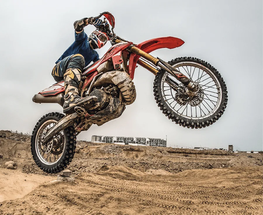 Photographie d'un pilote de motocross en plein saut, sur une moto rouge. L'action se déroule sur un terrain sablonneux.