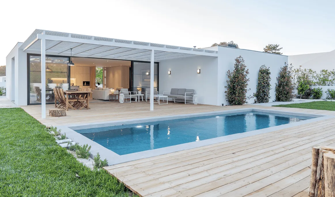 Photographie d'une piscine rectangulaire bleue, intégrée à une villa moderne blanche avec terrasse en bois.