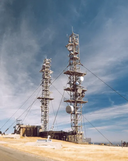 Photographie de deux pylônes de télécommunications équipés d'antennes, sur fond de ciel bleu et terrain sec.