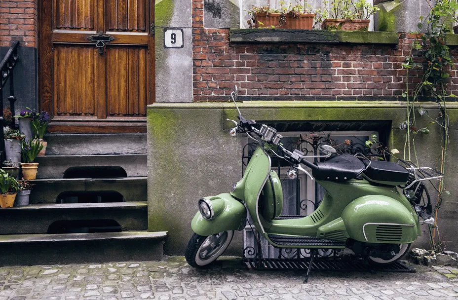 Photographie d'un scooter vert pâle garé devant une maison ancienne.