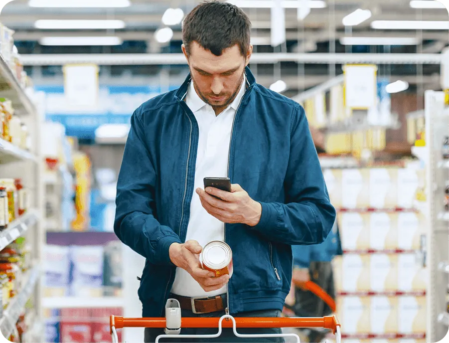 Photographie d'un homme dans un supermarché, consultant son téléphone portable et examinant une conserve.  Il utilise probablement une application pour comparer les produits.