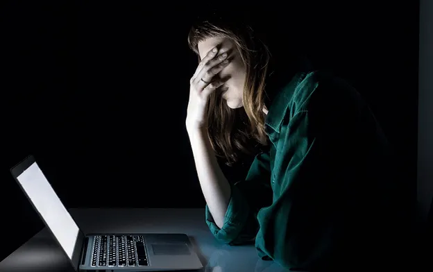 Photographie d'une femme fatiguée, la tête dans les mains, devant un ordinateur portable. Fatigue numérique, stress.